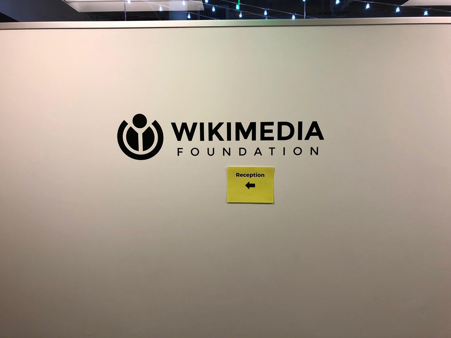 The Wikimedia Foundation name and official symbol on a wall at its former San Francisco office entrance, with a small reception sign below.