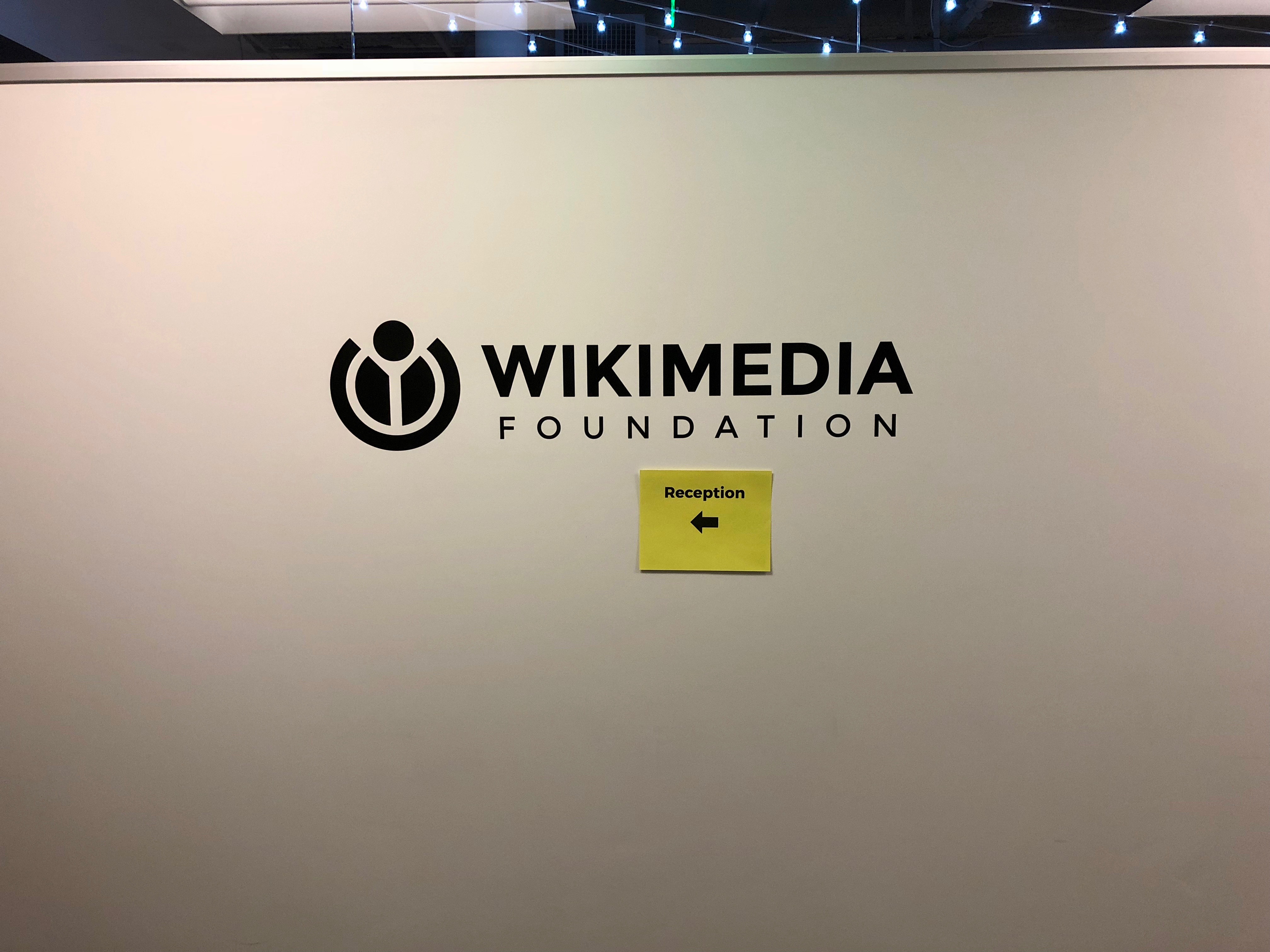 The Wikimedia Foundation name and official symbol on a wall at its former San Francisco office entrance, with a small reception sign below.